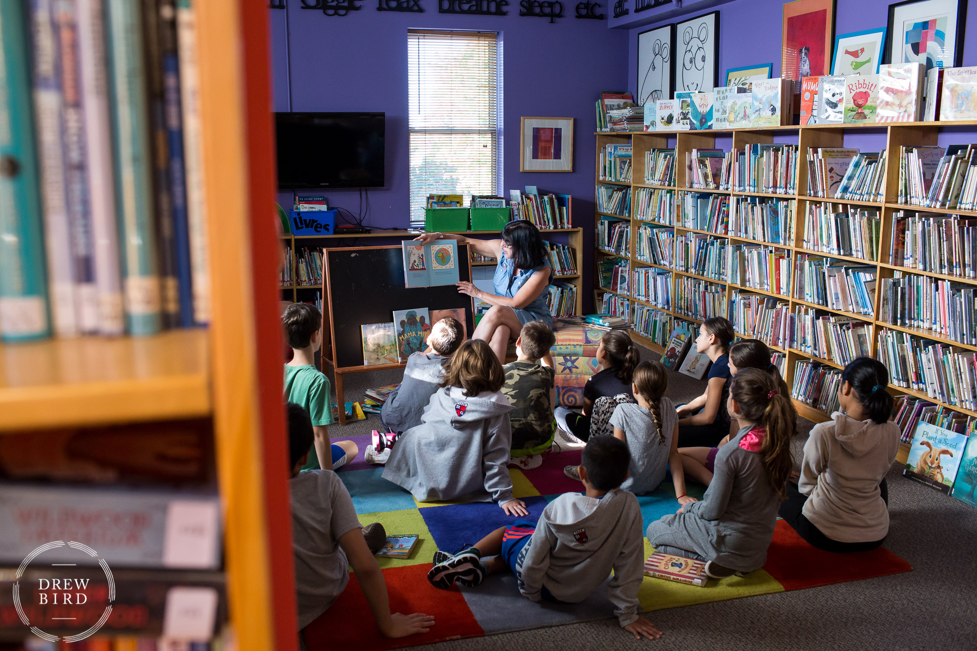 A woman teacher reads a book to a group of primary school students sitting on the floor in library at St. George's School of Montreal in Quebec, Canada. San Francisco independent school photographer and private school marketing photography and brand lifestyle photography by Drew Bird.